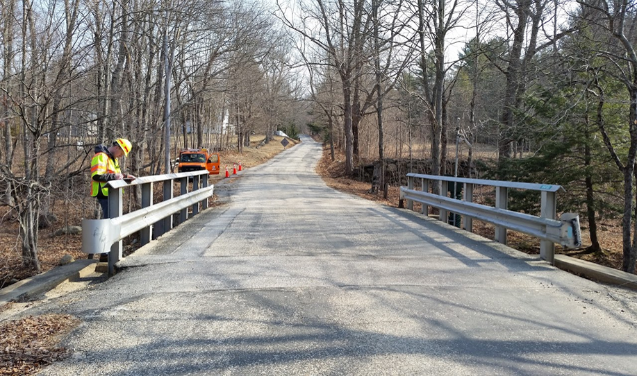 A bridge inspector in a vest and hard hat stands behind the bridge rail and inspects the travelway surface carried over the bridge. An inspection vehicle parked off the roadway and traffic cones are in the distance of an otherwise empty approach roadway.
